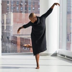 A person with a shaved head wearing a black dress performs a dance pose in a bright room with large windows. One leg is raised behind, and arms are outstretched gracefully. Sunlight streams in through the windows, illuminating the space and cityscape outside as they wear AirPods 2.