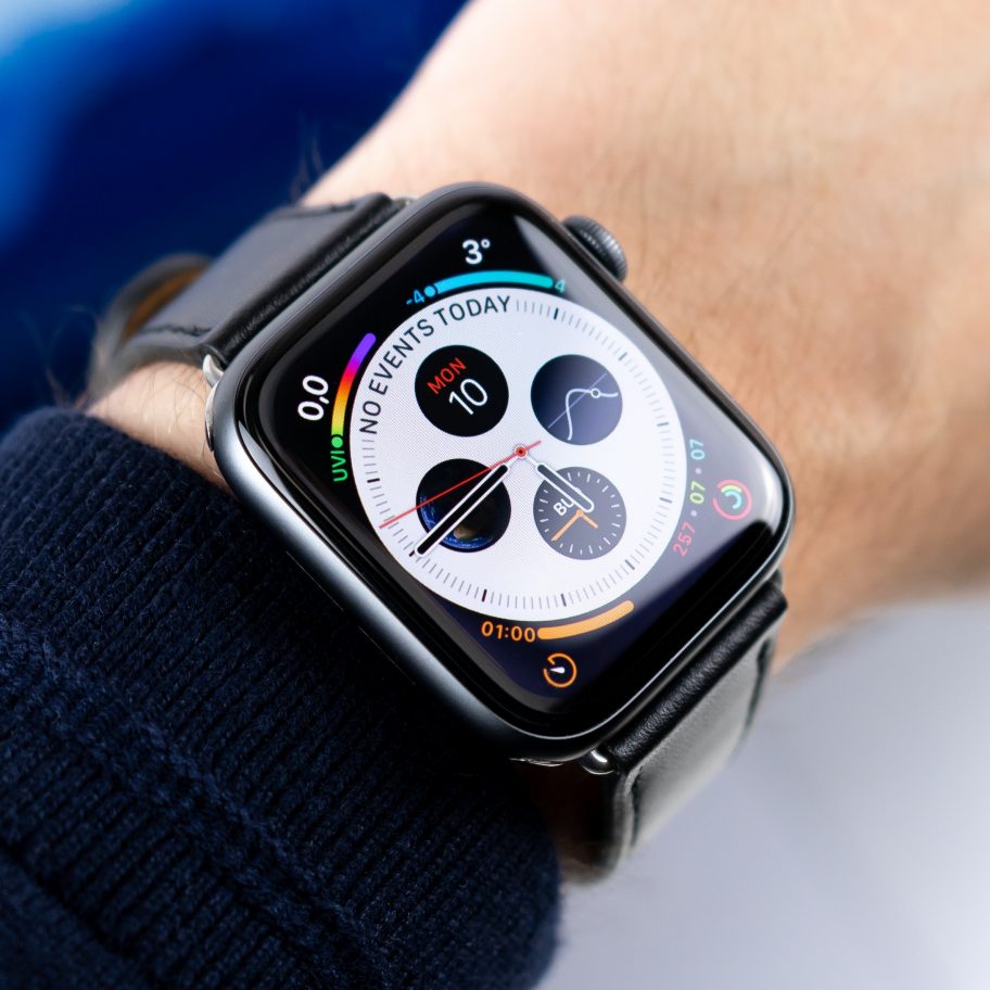 Close-up of a person's wrist wearing an Apple Watch 5 with a black band. The watch display showcases various Apple Watch concepts, including colorful activity rings, date, time, weather, a heart rate graph, and multiple widgets. The background is blurred blue and white.