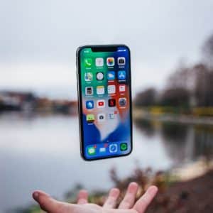 A person's hand is extended upward as an iPhone appears to levitate above it, showcasing various app icons. The background features an outdoor scene with a body of water and trees, indicating a park or riverside location. It's a perfect blend of iOS magic with nature's tranquility.