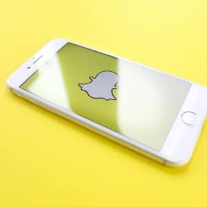 A white smartphone with the Snapchat app open is placed on a yellow surface. The screen displays the Snapchat ghost logo prominently. The phone is angled slightly, reminding users of past concerns about employees snooping on private messages.