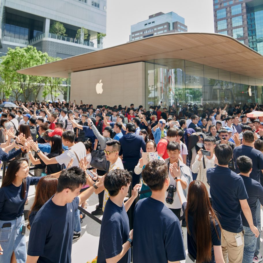 A large crowd gathers outside the latest Apple Store, Apple Xinyi A13, with many holding up phones to take pictures. The store has a modern glass and steel facade. Apple Store employees in navy blue shirts are lined up, forming a pathway and high-fiving visitors.