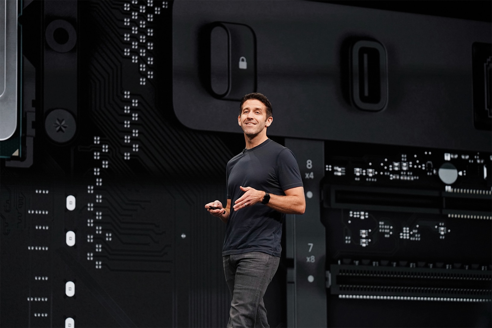 WWDC26 AI reveal - A person in a dark t-shirt and jeans stands on a stage, smiling and holding a device. Behind them, an enlarged image of a tech circuit board showcases various electronic components and connectors, reminiscent of everything we know about the Mac Pro’s advanced technology.