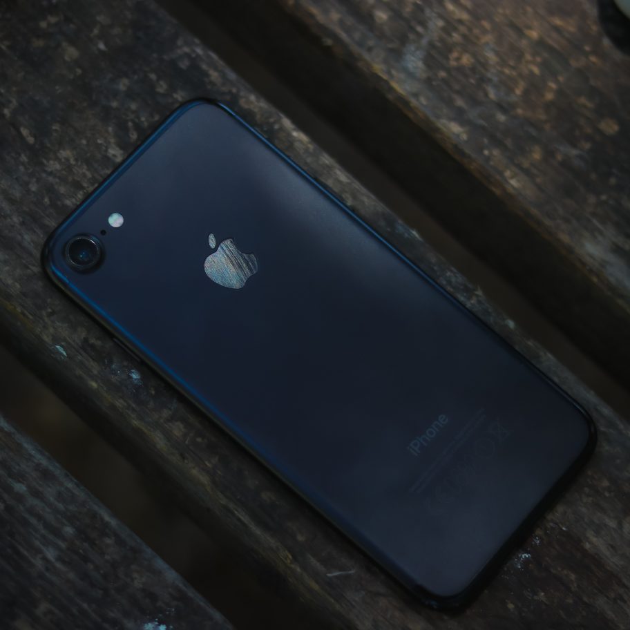 A black iPhone is placed face down on a wooden surface, with the Apple logo clearly visible. The lighting is dim, casting shadows on the phone and the wooden background, reminiscent of minimalist designs often seen in India.