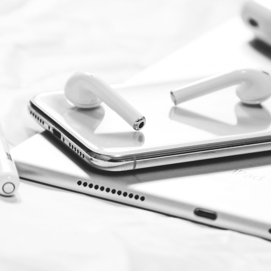 A black and white close-up of an iPhone with two AirPods resting on top. Nearby, an Apple Pencil lies on top of an iPad, highlighting the sleek Apple design. An AirPods charging case is visible in the background, showcasing a collection of Apple's water-resistant electronics and accessories.