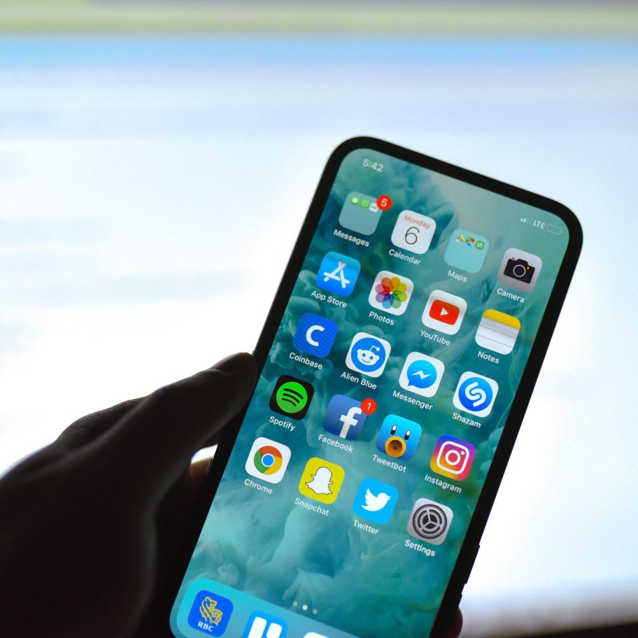 A person holds a notch-less iPhone displaying various app icons, including Messages, Calendar, Photos, YouTube, Facebook, Instagram, Spotify, Snapchat, Twitter, and others. The background shows a blurred computer screen. The person’s hand is slightly shadowed.