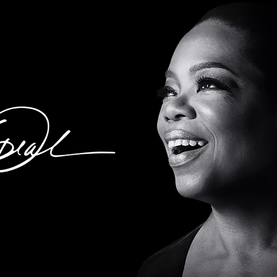 A black and white image of a woman smiling and looking to the right. She is well-lit against a dark background, and her facial features are clear. A cursive signature displayed beside her spells out "Oprah," reminiscent of classic shows now streaming on Apple TV+.