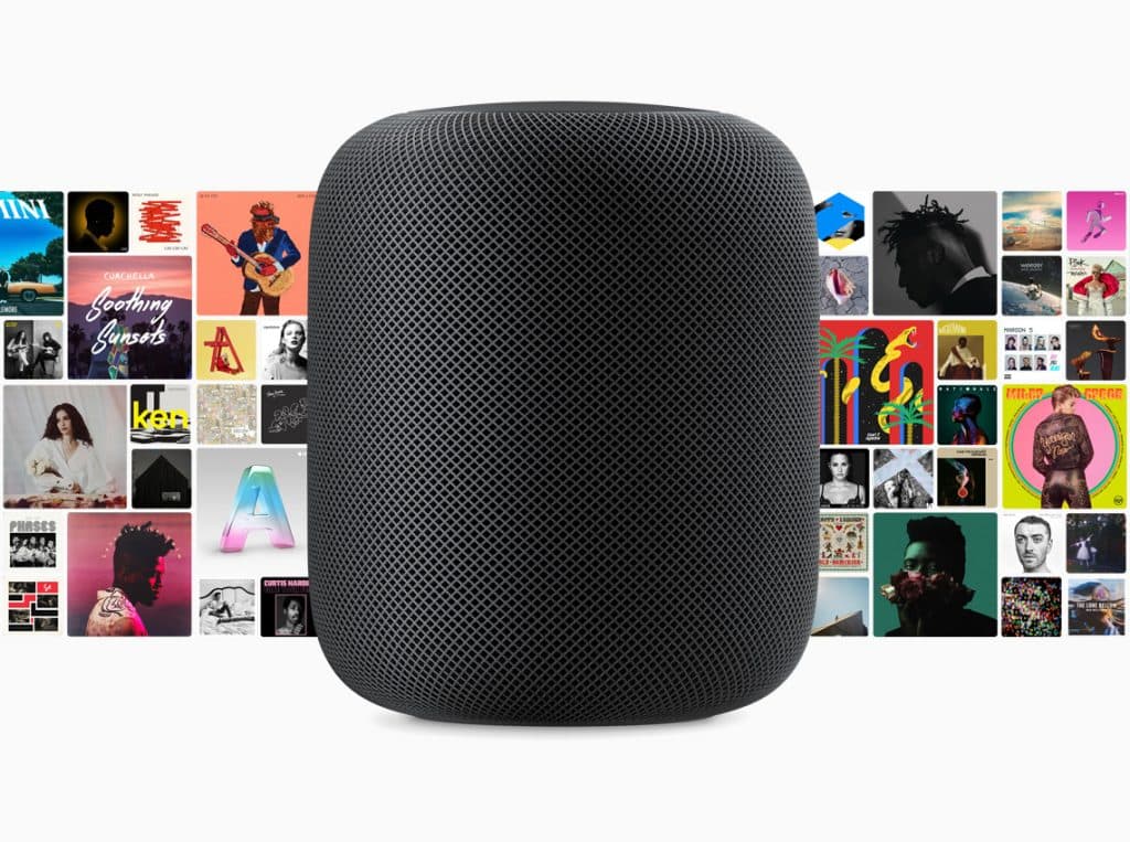 A black smart speaker is centered in front of a colorful collage of various album covers. The speaker, reminiscent of reveals at an Apple October Event, has a mesh fabric exterior and a cylindrical shape with a rounded top. The album covers feature diverse artwork and vibrant designs.