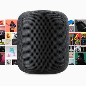 A black smart speaker is centered in front of a colorful collage of various album covers. The speaker, reminiscent of reveals at an Apple October Event, has a mesh fabric exterior and a cylindrical shape with a rounded top. The album covers feature diverse artwork and vibrant designs.