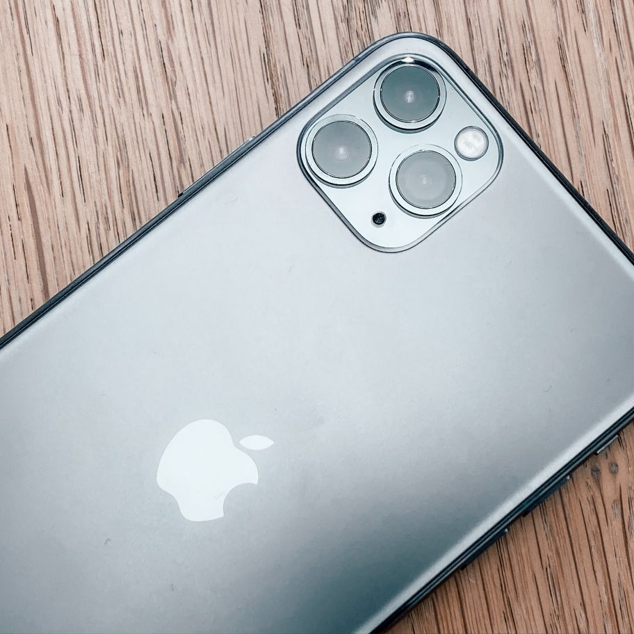 A close-up of a silver or gray iPhone lying on a wooden surface, showcasing the back of the device. The Apple logo is prominently displayed in the middle. The phone has a triple-camera setup in the top-left corner, consisting of three lenses and a flash, featuring higher sustained graphics performance thanks to its A13 Bionic chip.