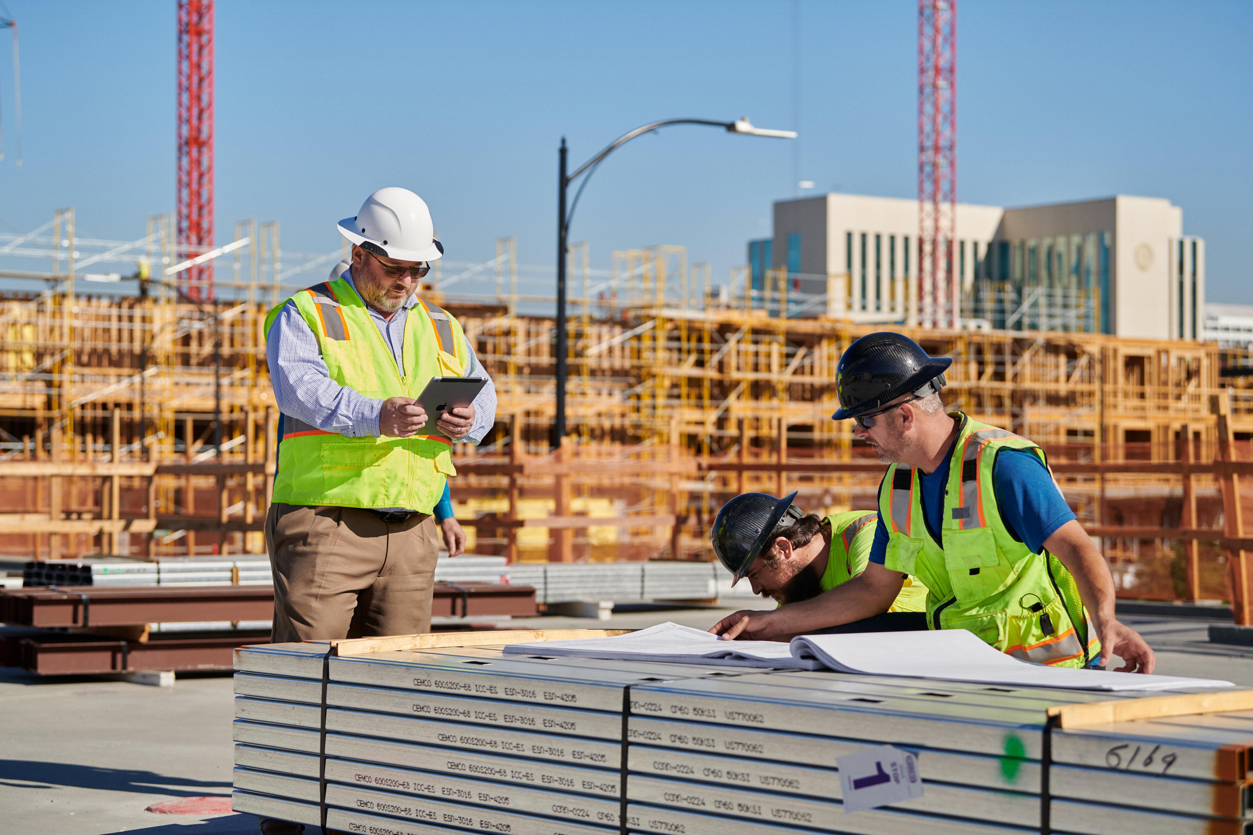 Three construction workers, wearing safety helmets and high-visibility vests, review building plans on a stack of materials at a construction site. The background shows partially constructed buildings, cranes, and a clear blue sky. One worker is using an Apple tablet, highlighting the $2.5 billion effort to tackle the California housing crisis.