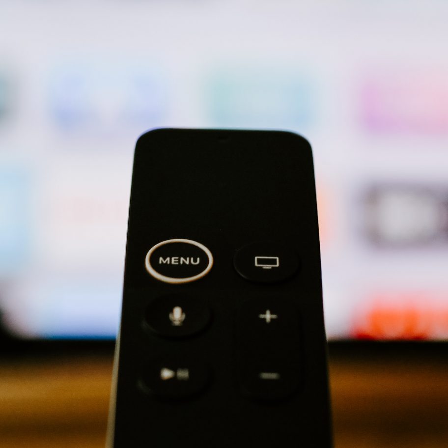 A close-up of a black remote control with several buttons, including a prominent "Menu" button, in front of a blurred TV screen displaying various apps and channels. The remote is centered and slightly out of focus, hinting at its compatibility with Apple TV for LG TVs coming months.