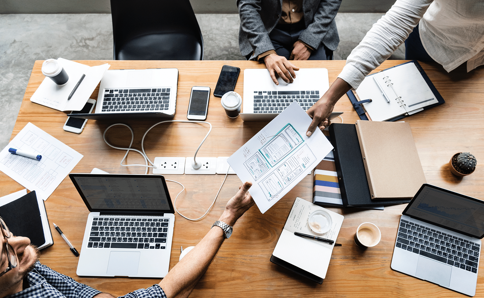 A group of people are seated around a wooden table at Apple Headquarters, each with a laptop. Papers, smartphones, coffee cups, notebooks, and other office supplies are scattered on the table. One person is handing a document to another. It's a collaborative work environment focused on DevOps strategies.