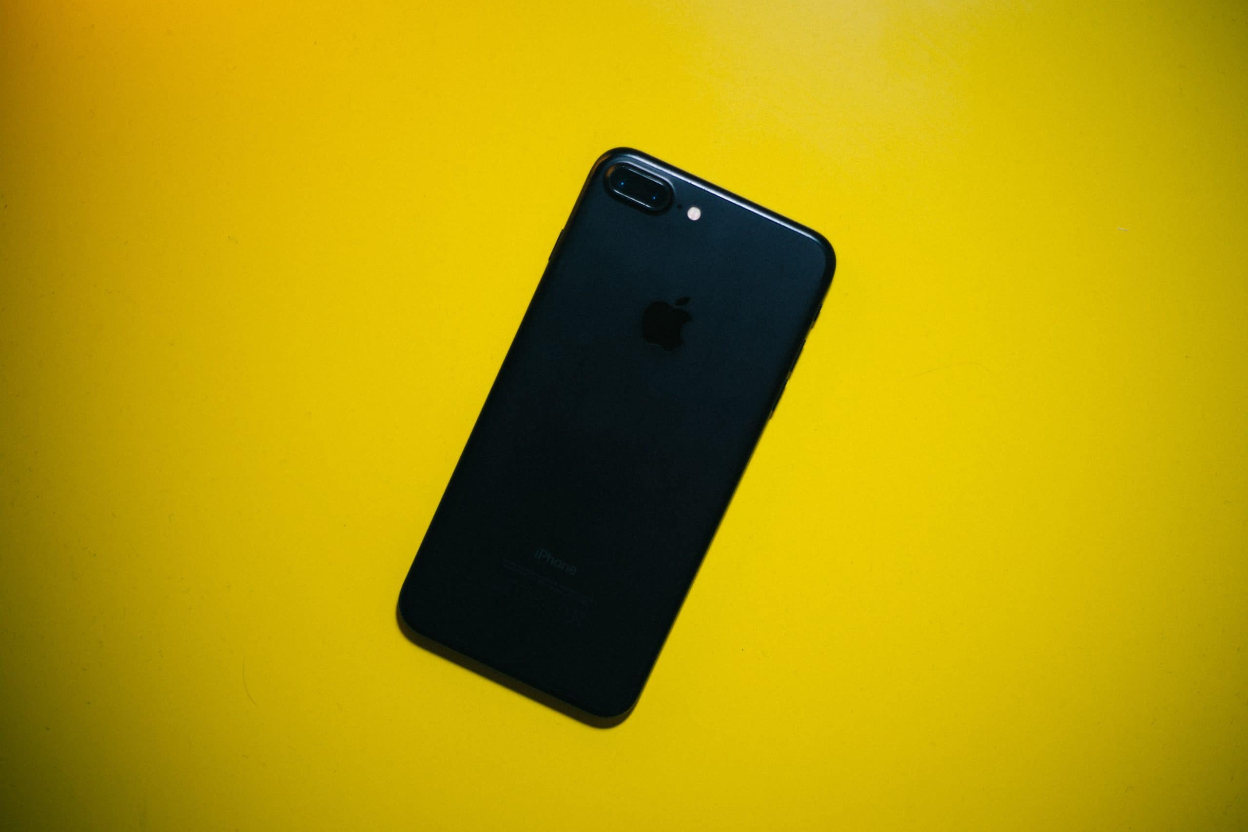 A black iPhone 7 lies face down on a bright yellow surface, showcasing the Apple logo and dual camera on the back.