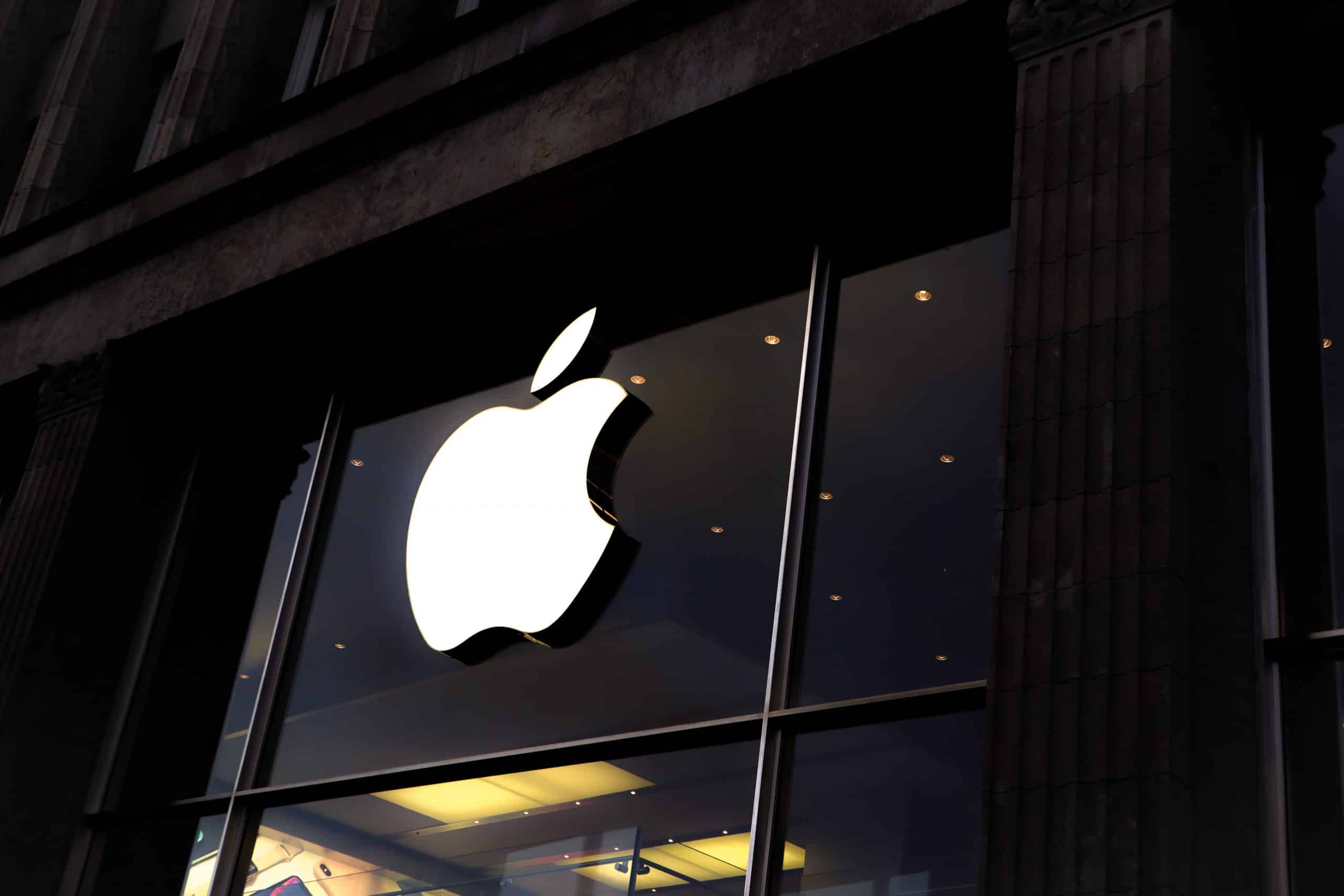 EU cloud scrutiny - A large, illuminated Apple logo is mounted on the exterior glass of a storefront. The logo is prominently displayed, with the interior of the store faintly visible through the glass. The building appears to be made of dark stone or concrete, highlighting the pros and cons discussed by customers considering AppleCare+.