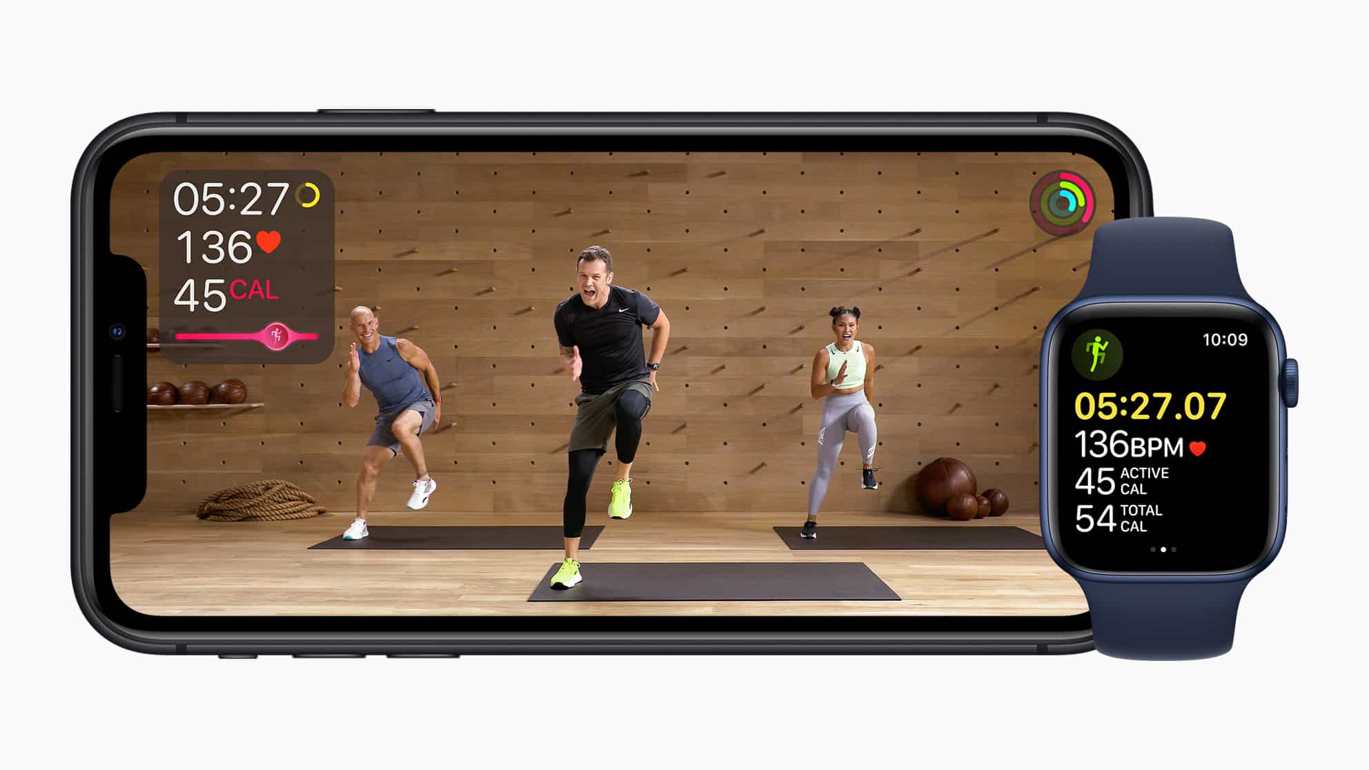 An iPhone displays a workout video with an instructor and two participants performing exercises in a wooden studio. Next to the iPhone, an Apple Watch shows matching workout metrics, including time, active calories burned, total calories, and heart rate.