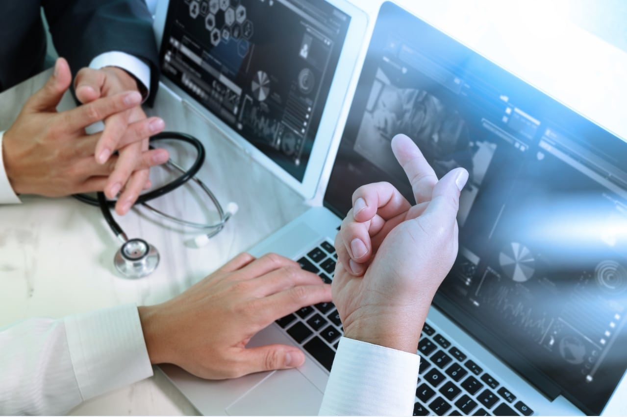 Two medical professionals discuss something displayed on a laptop. One person points at the screen, while the other uses a computer mouse. A stethoscope and another screen displaying medical charts are visible in the background, highlighting the evolving technology in the healthcare industry.