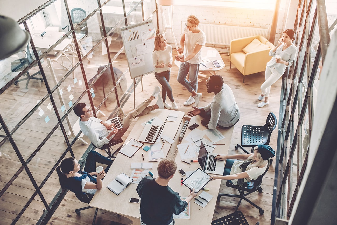 A diverse group of people is gathered in a modern office space with large windows. They are engaged in a collaborative meeting around a table filled with laptops, papers, and charts. Some are sitting, while others stand or lean against surfaces, discussing company changes and up-to-date solutions.