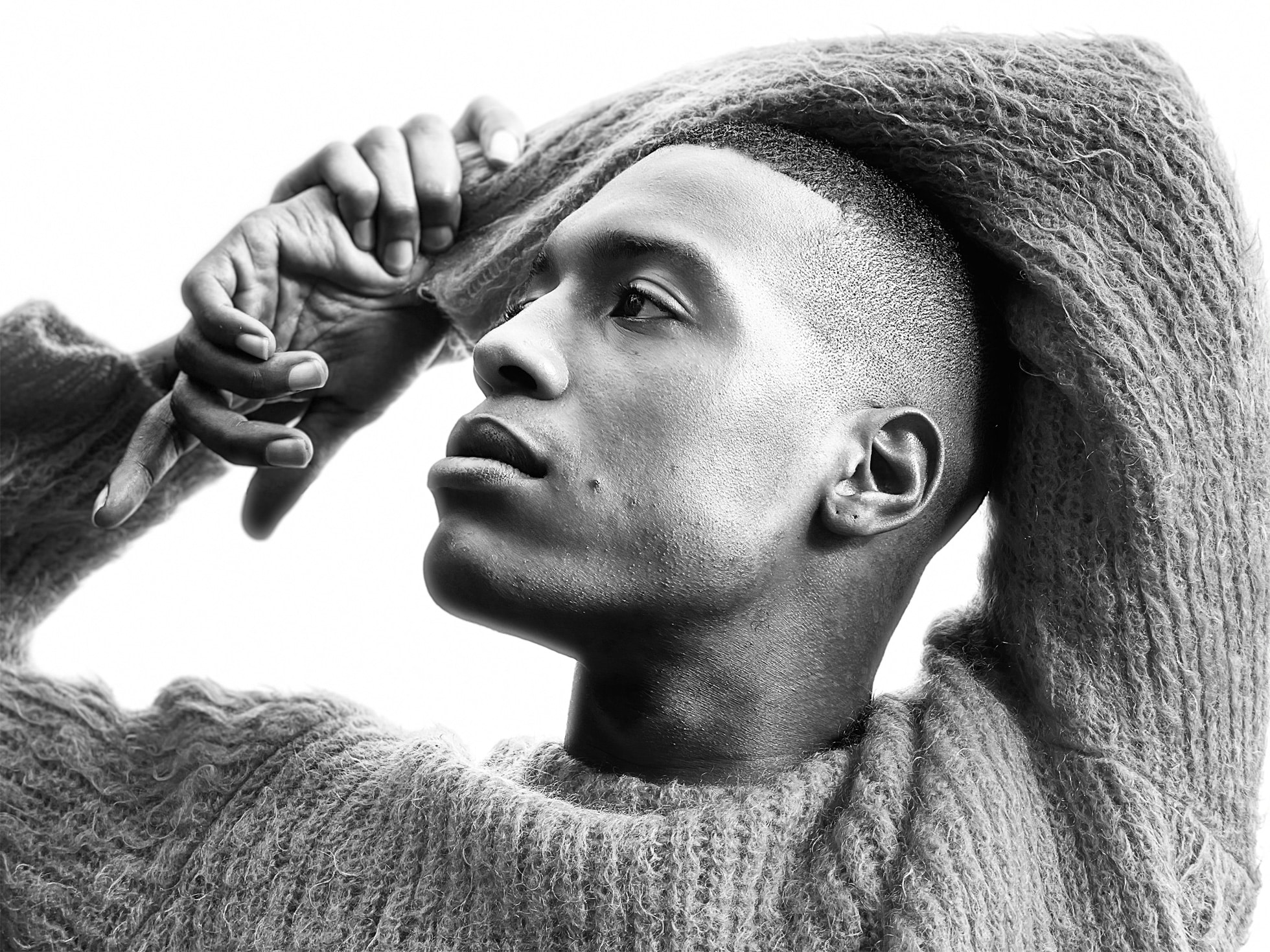 A black and white photo, likely shot with an iPhone 12 camera, captures a man in a textured knit sweater. He's shown in profile, lifting the collar over his head with one hand and tilting his head slightly back. His facial expression appears contemplative and calm against a plain background.