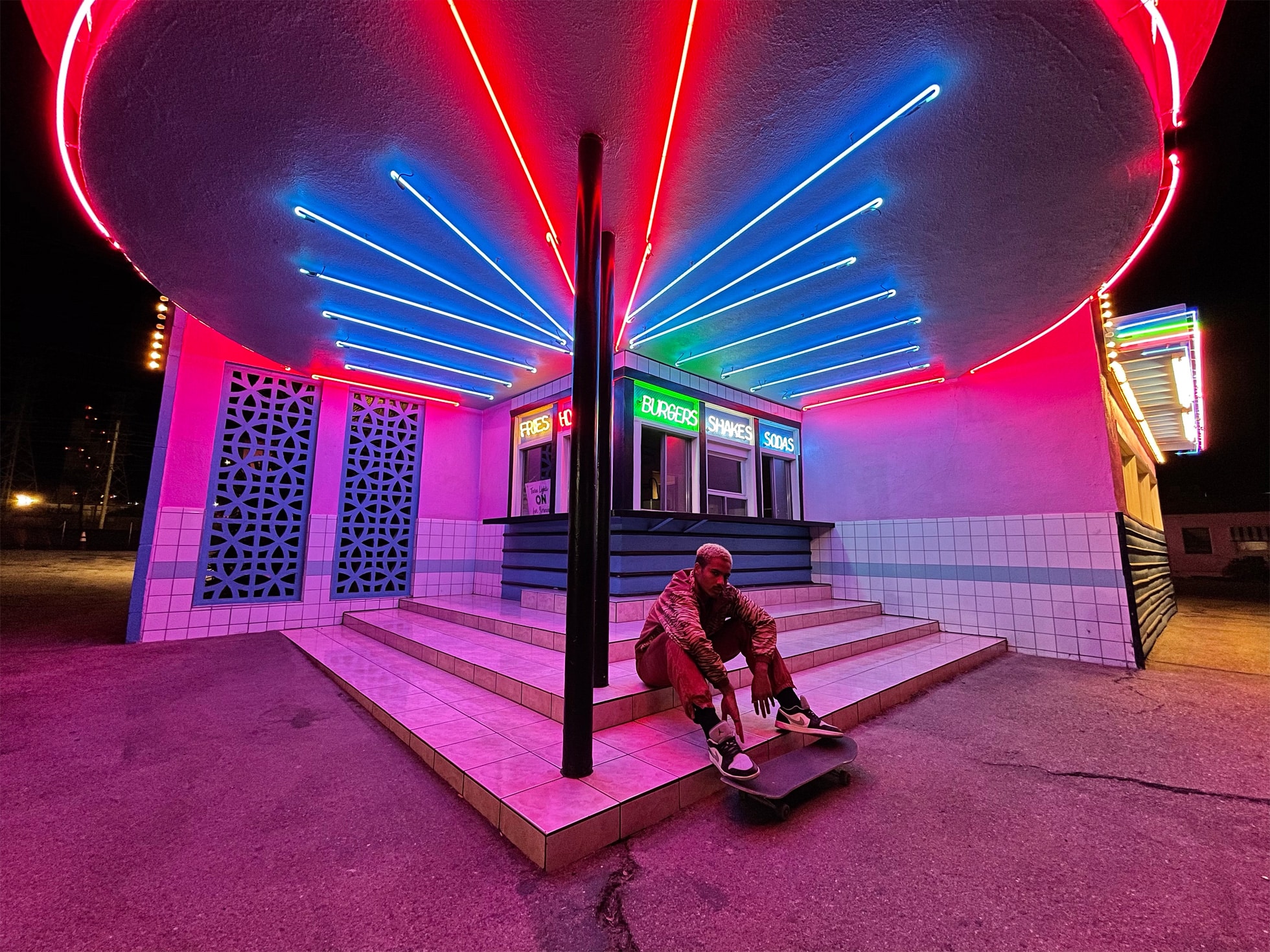 A person sits on a skateboard at the brightly lit entrance of a neon-lit diner at night, capturing the vibrant scene with an iPhone 12 camera. The neon signs above read "Fries," "Burgers," "Shakes," and "Sundaes." The diner's geometric lattice wall and tiled exterior add to the perfect shot for photography apps.