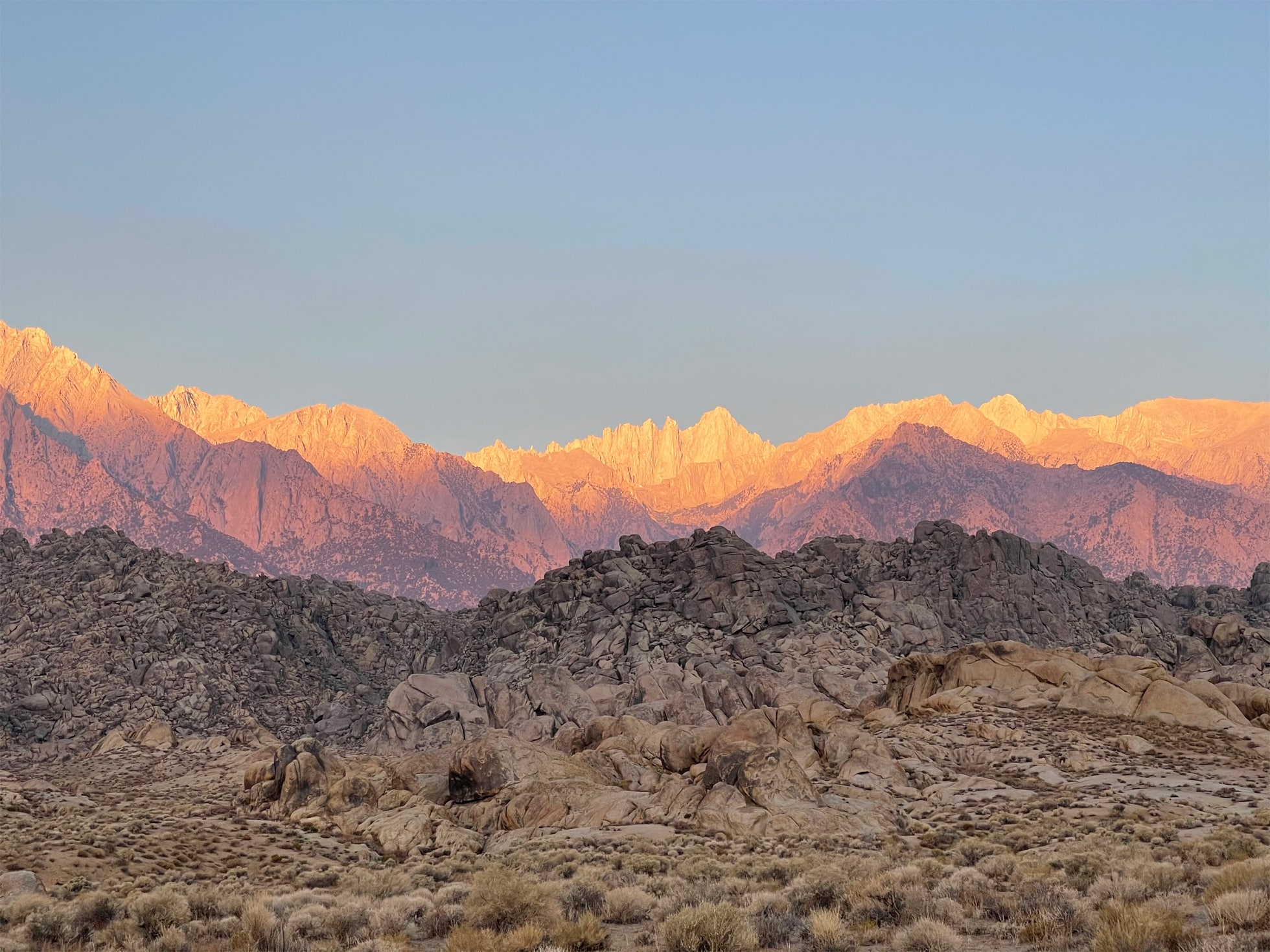 A panoramic view of a mountain range under a clear sky, with sunlit peaks displaying a warm, golden hue. Rocky formations and arid brush cover the foreground, transitioning to the rugged mountains in the background. The stunning detail captured by an iPhone 12 camera creates a striking contrast between light and shadow.