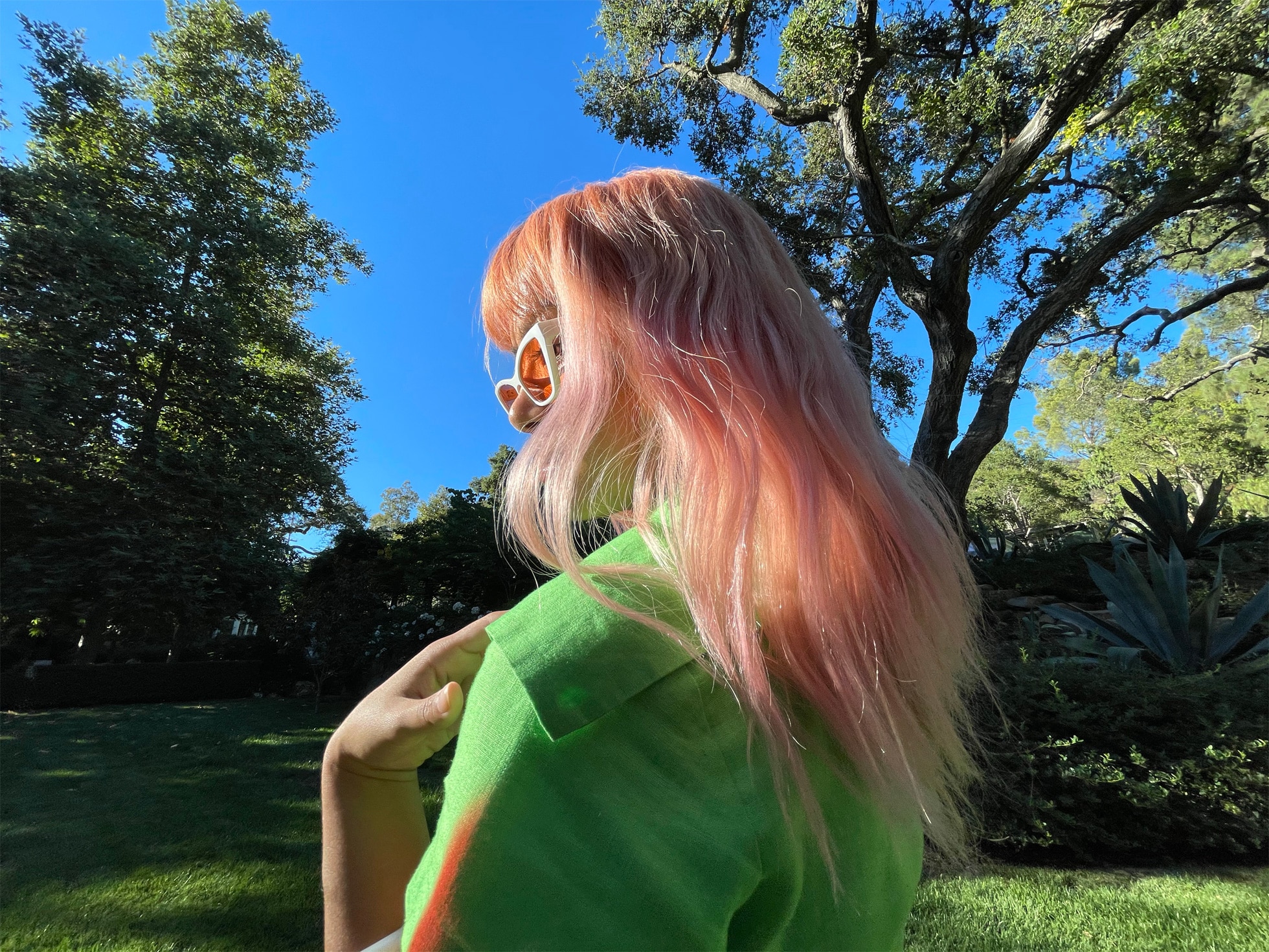 A person with long, pink hair stands outdoors in sunlight, wearing white sunglasses and a green shirt. They are turned away from the camera, showcasing their hair. Captured with an iPhone 12 camera, the background features lush green trees and a clear blue sky—truly taking your photography further.