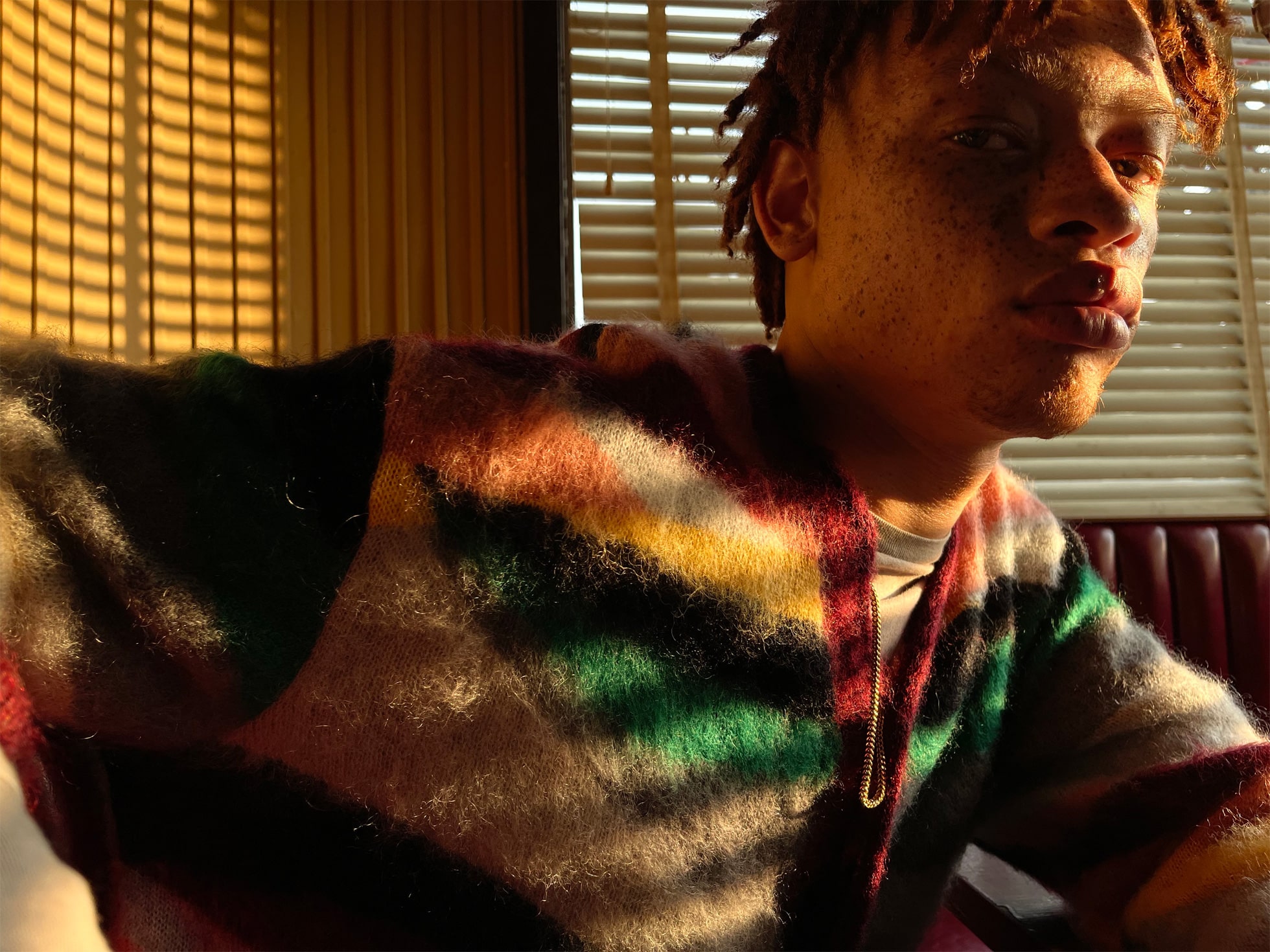 A person with short red hair and freckles sits in a room with blinds, an iPhone 12 in hand. Warm sunlight creates patterns of light and shadow on their face and multicolored striped sweater. They gaze pensively to the side, with a serious expression, as if contemplating the perfect shot for their photography app.