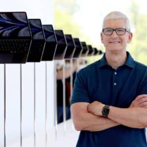 Tim Cook stands with arms crossed in front of a lineup of MacBook laptops displayed on stands during an Apple event.