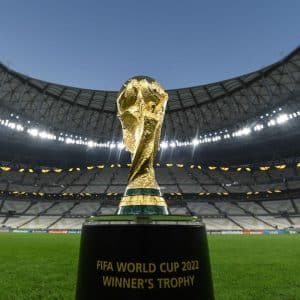Image of the FIFA World Cup 2022 Winner's Trophy displayed in an empty stadium. The gold trophy stands prominently on a black pedestal against the backdrop of the stadium's seating and roof structure, showcasing its intricate design under stadium lights, akin to those seen during FIFA Club World Cup broadcasts.