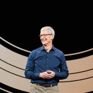 A person with short gray hair and glasses stands on a stage, smiling while wearing a dark blue shirt and khaki pants. The background features large, curved, concentric lines, setting the scene for the anticipated discussion on Q2 2024 analyst expectations for Apple earnings.