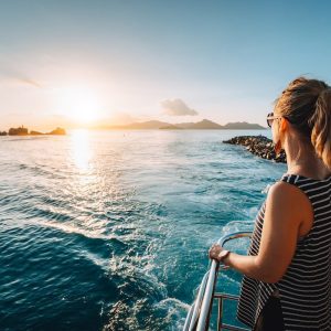 A woman with blonde hair in a ponytail stands on the deck of a boat, gazing at a beautiful sunset over the ocean. She wears a sleeveless striped top and sunglasses. The water sparkles in the sunlight, and distant mountains and other boats are visible on the horizon. Capturing this moment would be a breeze with iOS 18's new camera features.
