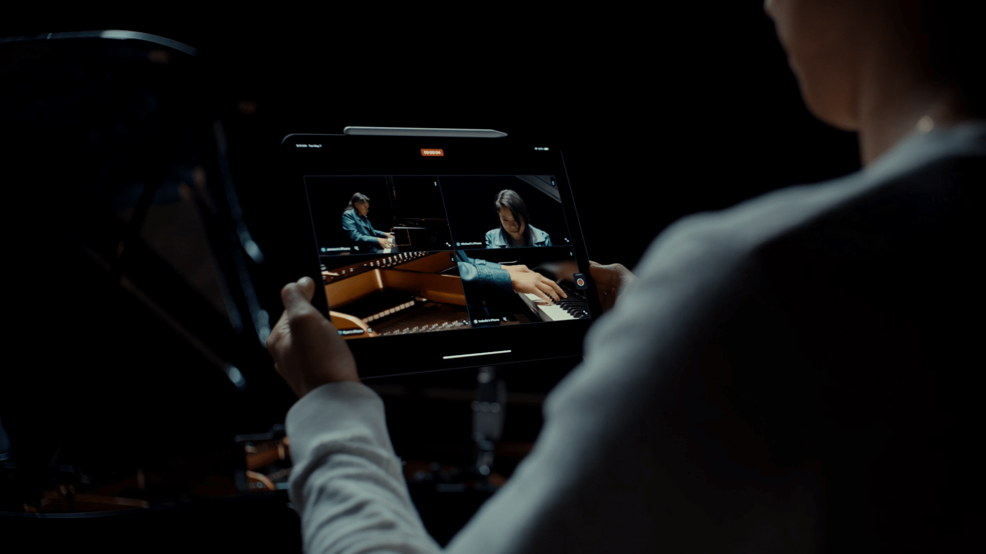 A person holds an iPad displaying a video with four split-screen views: an overhead shot, a side view, a close-up of hands on a piano, and a person playing the piano. The background is dark, highlighting the bright screen of the tablet showcasing the impressive Multicam Feature in Final Cut Pro.