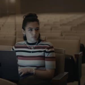 A woman with a striped shirt sits alone in an empty auditorium, working on a laptop. She has dark hair tied up and wears multiple necklaces and bracelets. The dim lighting highlights her focused expression as she benefits from impressive Mac battery life.