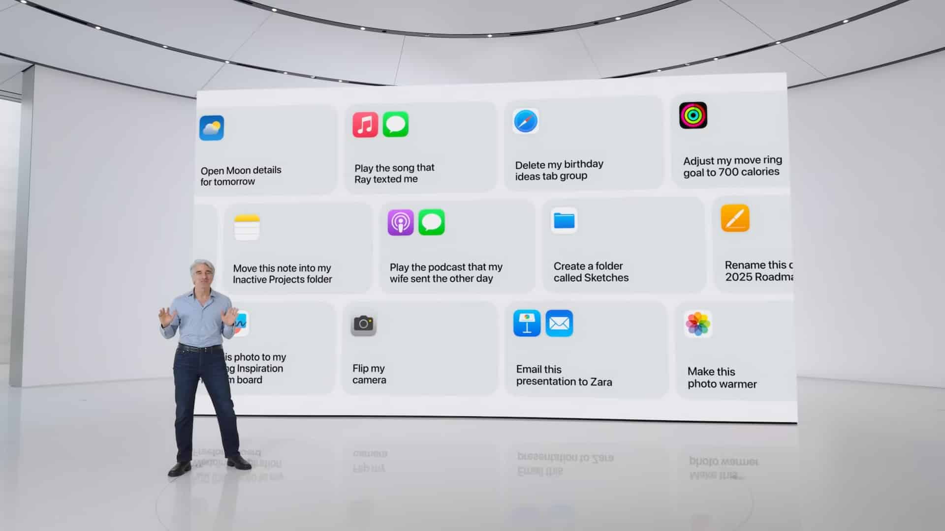 A speaker stands on stage gesturing towards a large screen displaying various app icons and their tasks, such as music, messaging, fitness, and camera controls. The background is minimalist and white, emphasizing the colorful app icons on the screen as they reveal what to expect from upcoming features.