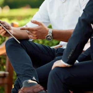 Two people are sitting on a bench outdoors, one wearing a white shirt and showing something on a tablet to the other, who is dressed in a dark suit. Their faces are not visible, but they appear to be engaged in a discussion about Google Ads strategy.