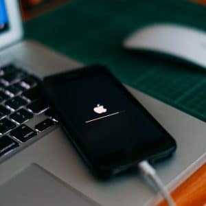 A smartphone displaying the Apple logo with a loading bar, potentially updating to iOS 18, is connected to a laptop with a grey keyboard. The laptop rests on a green cutting mat, and a white computer mouse is placed nearby on the wooden surface.