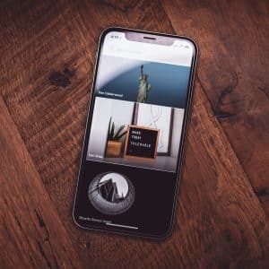 A sleek Apple smartphone displaying a gallery app lies on a wooden surface. The screen shows three photos: the Statue of Liberty, a framed quote saying "MAKE TODAY TOLERABLE" near some plants, and a black and white image of modern architecture, as if from a carefully recovered album.