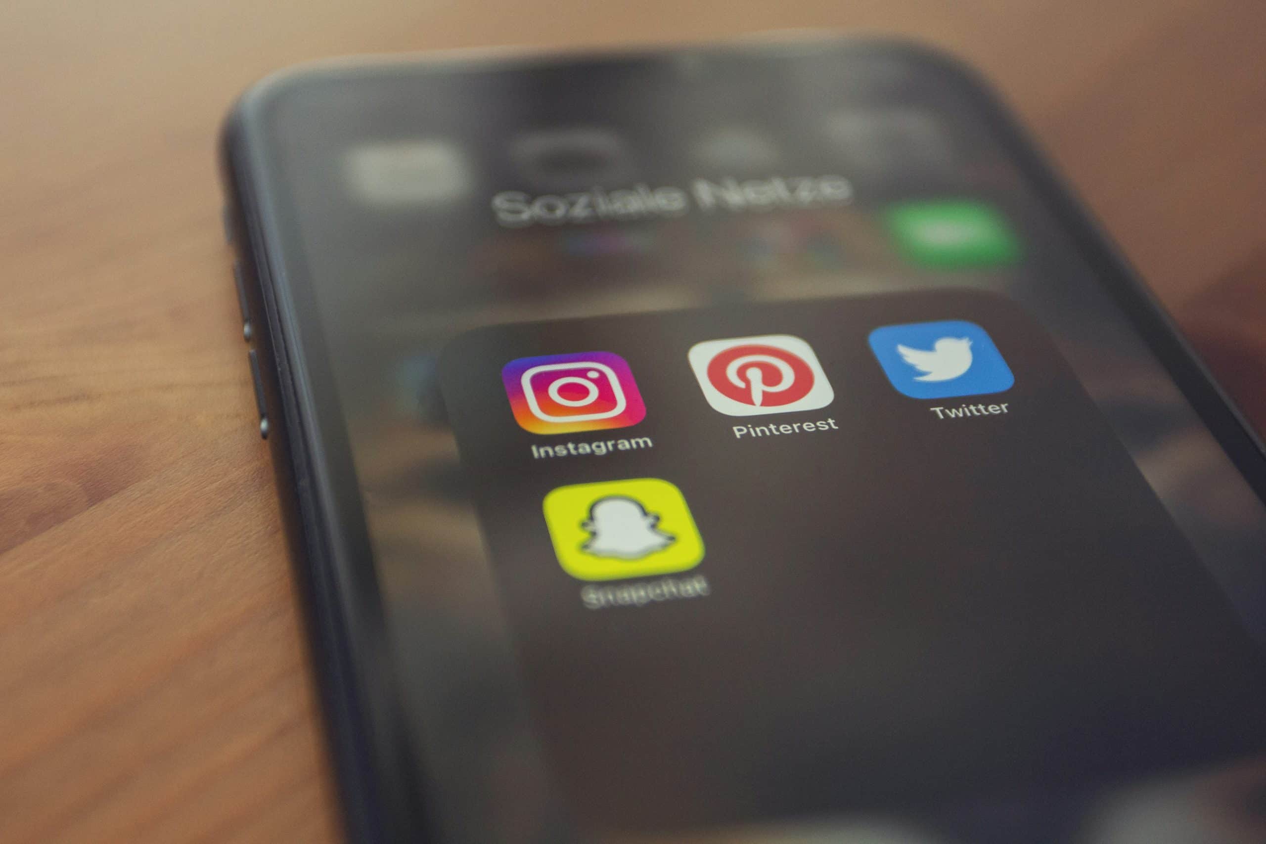 A smartphone screen displays a folder labeled "Soziale Netze" containing the icons for Instagram, Pinterest, Twitter, and Snapchat. The phone rests on a wooden surface, with Snapchat's icon standing out brightly among the others.