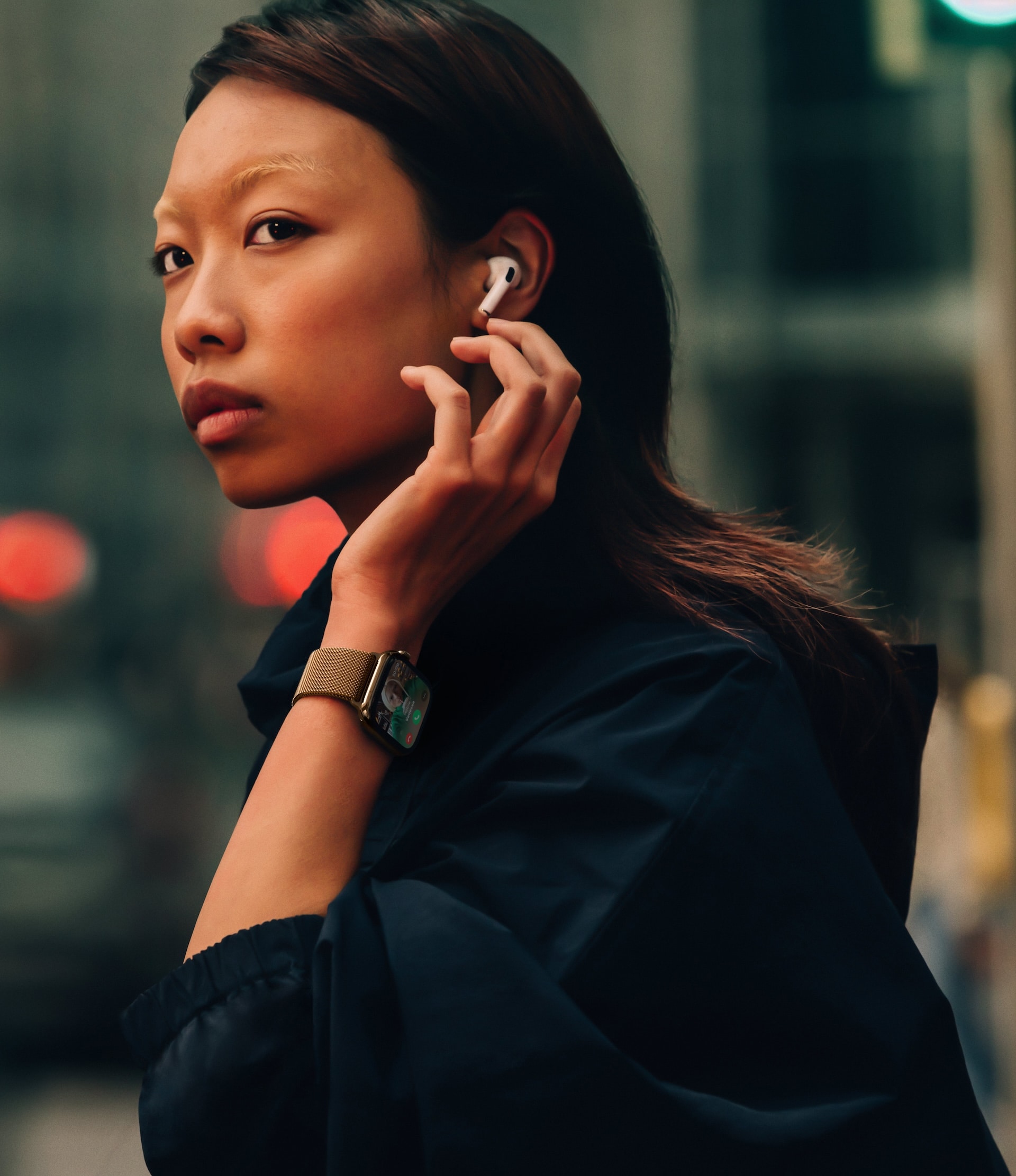 Apple Watch Series 10: A person with long hair, wearing an Apple Watch 10 and a dark outfit, inserts an earbud into their ear while looking over their shoulder on a city street. The background is blurred, with red and green lights visible.