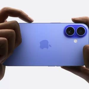 A person holds a blue iPhone 16 horizontally with both hands, focusing on its dual rear cameras. The AppleMagazine logo is visible in the center of the device.