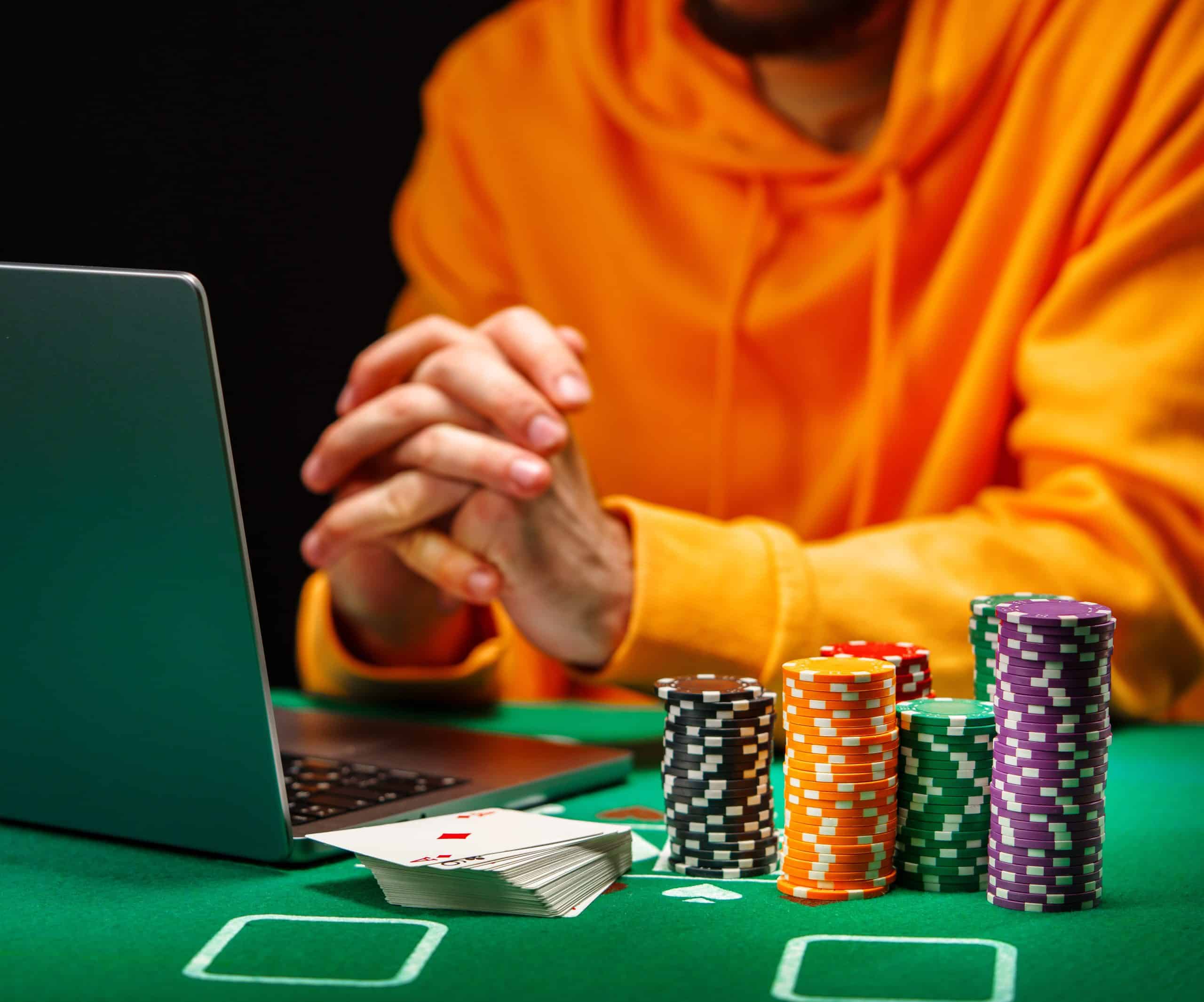 A person in a yellow hoodie sits at a table with a laptop, stacks of poker chips, and a spread of playing cards showing an ace of hearts. Their hands are folded in front of them, focusing intently.