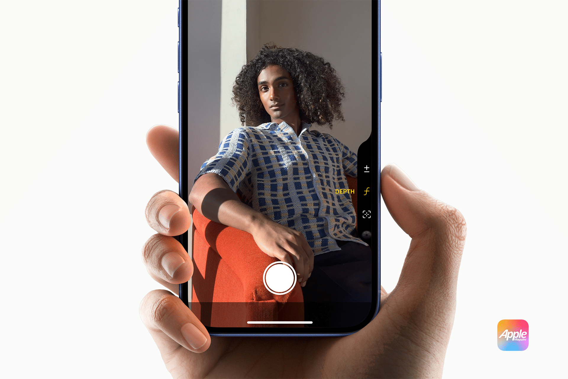 Close-up of a hand holding an iPhone 16, capturing a person seated elegantly on an orange chair. The phone screen displays the subject with camera settings visible. The white background and distinctive Apple logo hint at the recent sales surge of Apple's latest device.