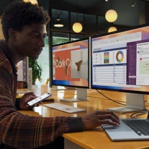 A person works at a desk in an office, focusing on their M4 MacBook Air with new colors. They hold a smartphone while two monitors display charts and a presentation. Another person in the background is also engrossed in their computer. The setting is modern and collaborative, echoing Apple's innovative spirit.