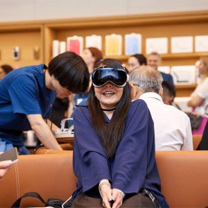 A person wearing Vision Pro VR goggles is sitting excitedly on a bench. They're surrounded by people in a brightly lit store, with electronics and phone cases displayed on the walls in the background.