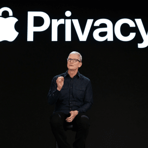 A person in dark clothing sits on a stool, gesturing while speaking on stage in front of a large screen displaying the Apple logo and the word "Privacy," highlighting iPhone security features.