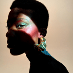A person with short hair poses in dramatic lighting, their iPhone 17 Pro capturing the play of shadows across their face. They wear bright makeup with shimmering colors and large, decorative earrings. The neutral background highlights the striking contrast of light and shadows.