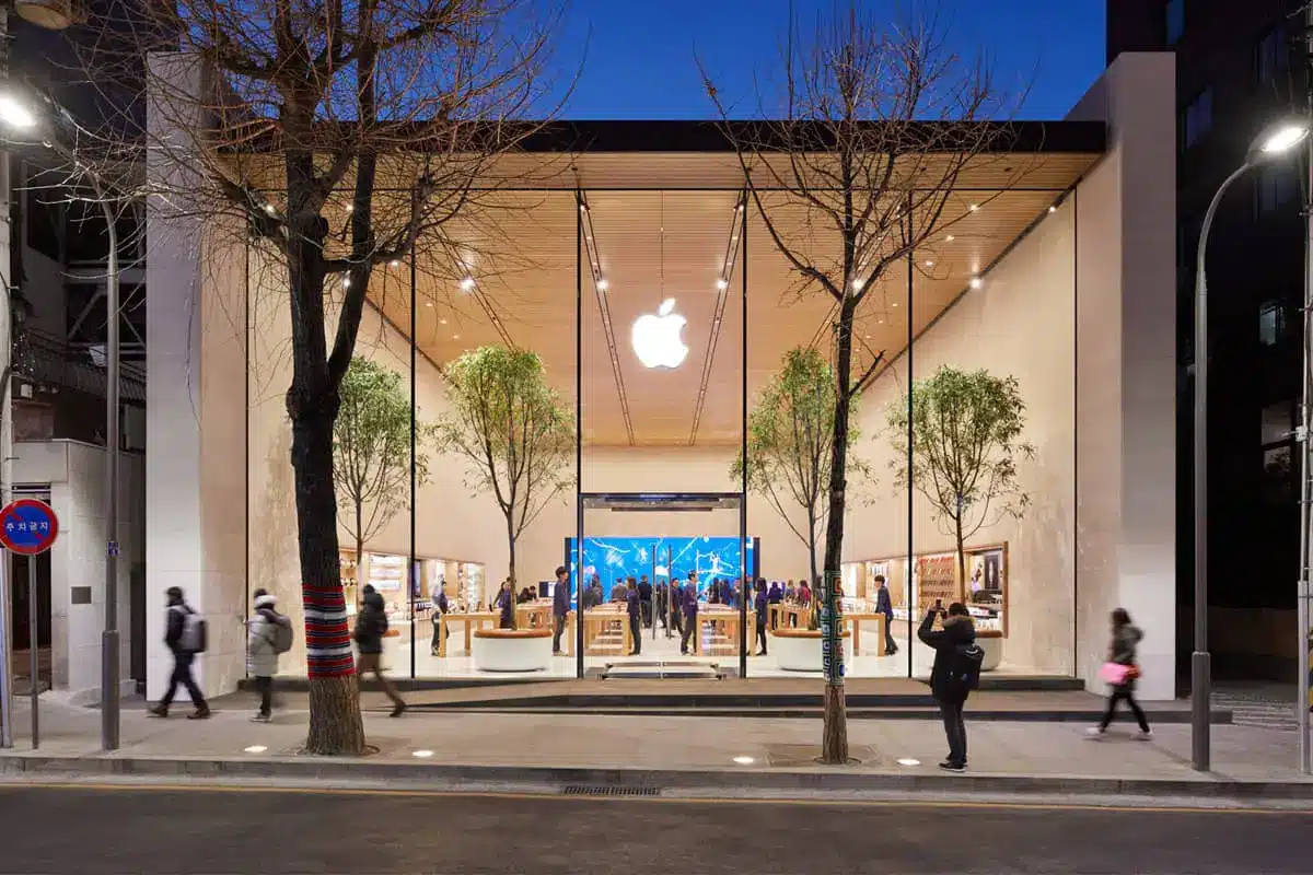 Apple options - A modern Apple Store with a glass facade, large Apple logo, and indoor trees draws crowds sightseeing in the city. People walk by or enter the brightly lit store during dusk, while the sky is deep blue.