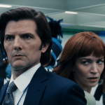 A man in a suit and a woman with red hair stand close together, looking in opposite directions with serious expressions—evoking the tense atmosphere of Severance—as people in blue and white uniforms move through the brightly lit room.