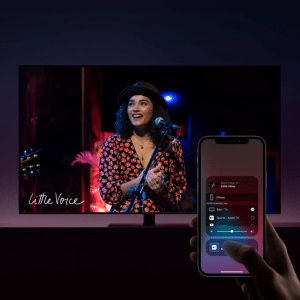 A person uses AirPlay on their smartphone to control media playback while a woman in a hat and red-patterned shirt performs on stage on a TV screen displaying "Little Voice.