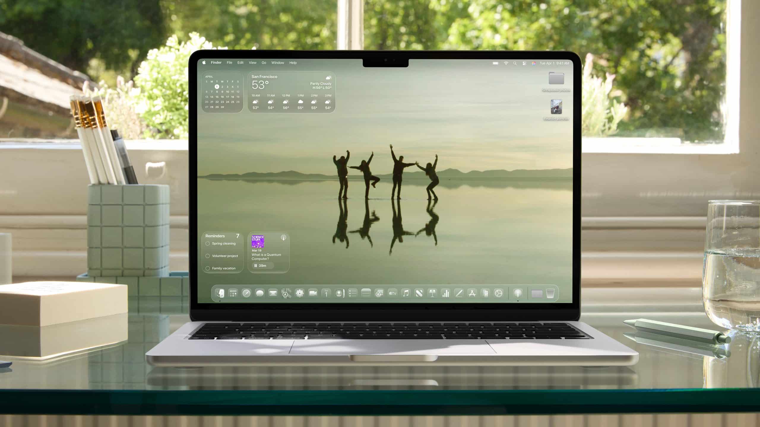 A laptop featuring Apple Intelligence sits on a glass desk, displaying a scenic wallpaper of four people dancing on a reflective surface. The desktop shows weather and widgets, with trees and sunlight visible through the window behind.