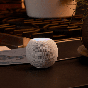 A white Apple smart speaker, possibly the HomePod Mini 2 (2025), with a glowing top sits on a kitchen counter near a stove, beside a potted plant and some papers.